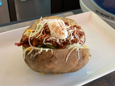 Baked potato split open, topped with brisket, cheese, bacon bits, sour cream, green onions on a white plate.