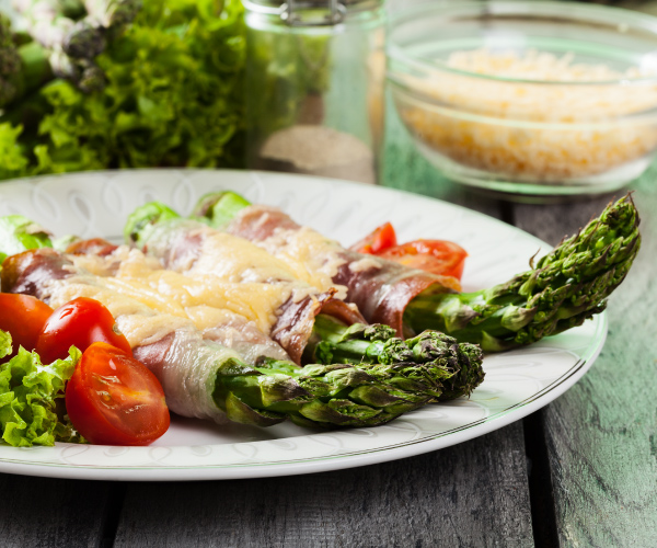 Finished asparagus on a round white plate sitting on a wood table, with a bowl of shredded cheese in the background.