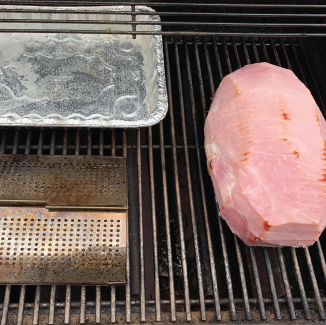 A whole ham sitting on a standup propane grill's cooking grid, with a smoking tray and aluminum pan with water in it.