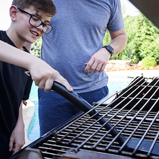 Father and son in the backyard standing in front of an open standup gas grill. Son is cleaning the cooking grates.