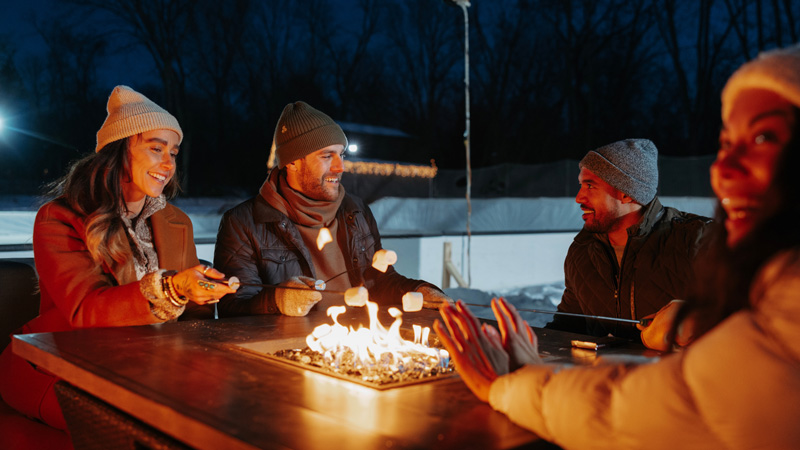 A group of friends in winter attire sitting outside at a propane powered fire pit laughing and roasting marshmallows.