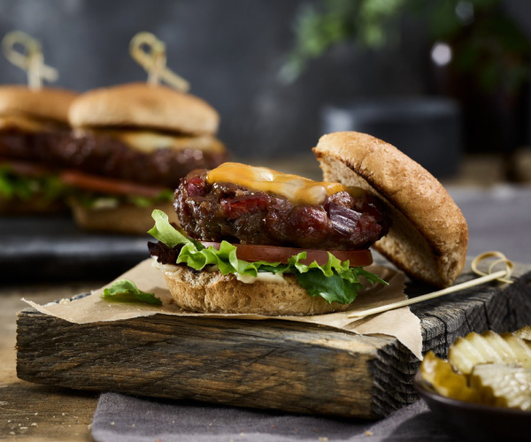 Beef Jerky slider on a small bun with cheese, lettuce and tomato on a wooden board with pickles on the side.