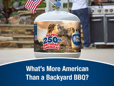 A fresh Blue Rhino propane tank with a special summer 2026 sleeve, sitting on concrete patio, with a man standing at standup propane grill in the background.