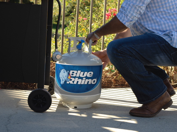 A man on a patio kneeling down by the side of a standup propane grill, getting ready to attach a fresh Blue Rhino proapne tank.