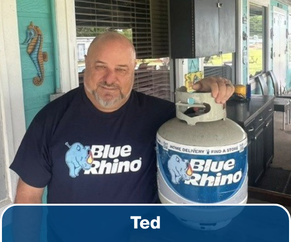 A man in a blue shirt standing beside a table with his arm on a Blue Rhino propane tank.