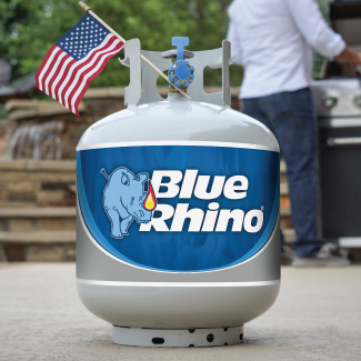 A fresh Blue Rhino propane tank with an American Flag in the collar, sitting on a patio, with a man grilling in the background.