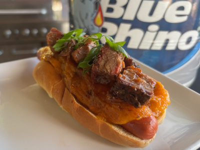 Finished bacon cheese dog on a white plate, with a fresh Blue Rhino propane tank and standup propane grill in the background.