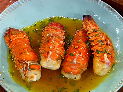 Four grilled lobster tails in butter mixture in a light blue bowl.