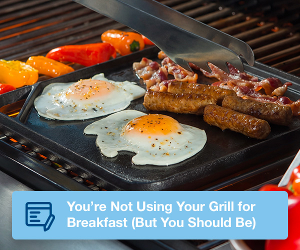 Eggs, sausage, and bacon being grilled on a cast-iron griddle topper on the cooking grids of a stand up propane grill.