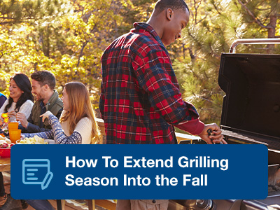 A man in a flannel shirt grilling at a standup propane grill, with friends at a patio table in the background.