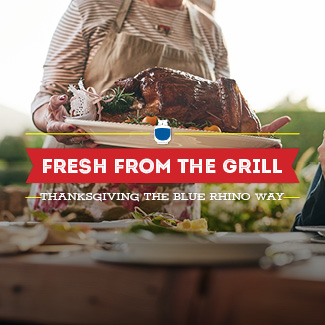 A woman in a tan apron carrying a finished grilled turkey on a white plate toward a dinner table.