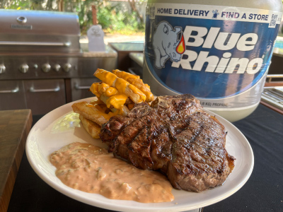 Finished steak frites with animal sauce on cheesy fries on a white plate, with a fresh Blue Rhino tank and standup propane grill in the background.
