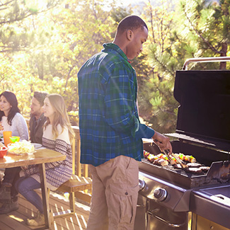 A man in fall attire grilling food on a standup propane grill, with friends sitting at a patio table in the background.