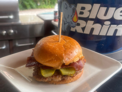 Finished Pork Smashburger on a white plate, sitting on a wooden cutting board, with a fresh Blue Rhino and a standup propane grill in the background.