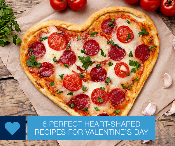 Finished heart-shaped pizza with pepperoni and sliced tomatoes sitting on a sheet of parchment paper on a wooden table.