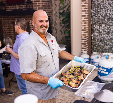 Chef holding an aluminum tray filled with grilled chicken, with fresh Blue Rhino propane tanks in the background.