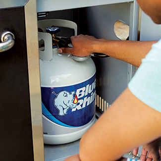 Closeup of a man connecting a standup propane grill's regulator to a fresh Blue Rhino tank.