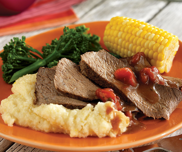 Slices of roast with sauce on bed of grits, with broccoli and grilled corn on an orange plate.