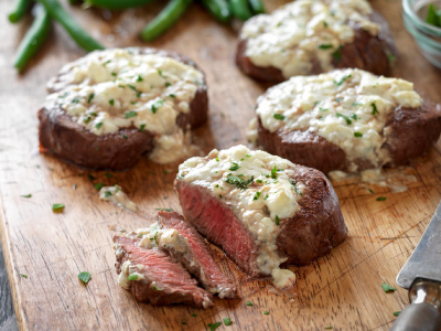 Four grilled steaks with blue cheese topping sitting on a wood cutting board.