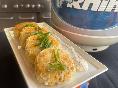 Finished crab cakes on a white plate, with a fresh Blue Rhino propane tank and standup propane grill in the background.