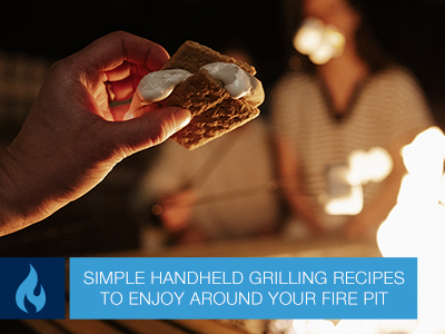 Mans hand holding a finished s'more with a gas fire pit and people in the background.