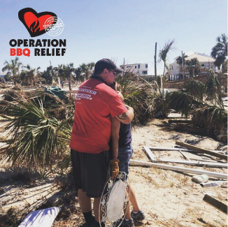 An Operation BBQ Relief volunteer hugging a hurricane victim, standing on sand, surrounded by downed palm trees and debris.