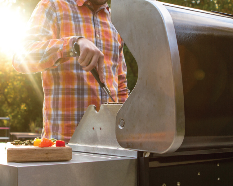 Man with plaid long sleeve shirt standing in front of stand up propane grill, using utensil to move food.
