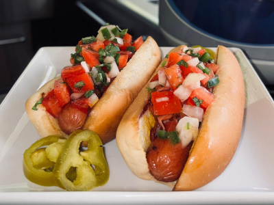 Two finished LA Street Dogs on a white plate, with a fresh Blue Rhino and standup propane grill in the background.