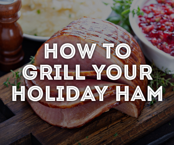 Finished smoked ham, sliced and sitting on a wood plate, with a bowl of cranberry sauce in the background.