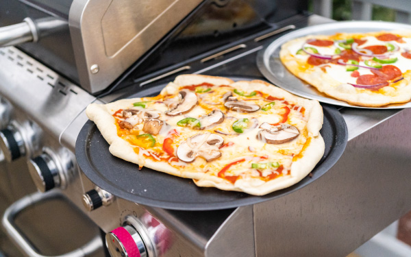 Two pizzas on pizza pans on standup propane grill's side shelf.