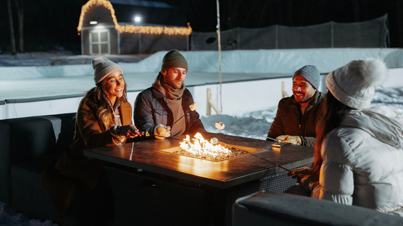 A group of friends sitting around a propane fire pit in the winter, with a hockey rink in the background.