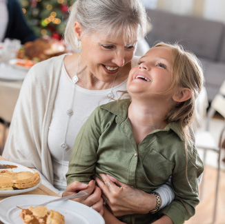 Grandmother holding the granddaughter on her lap, sitting at a wooden dining table with slices of apple pies on white plates.