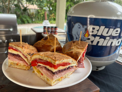 Finished sliced Crispy Muffuletta Sandwich on a white plate, on a wooden cutting board, with a fresh Blue Rhino and standup propane grill in the background.