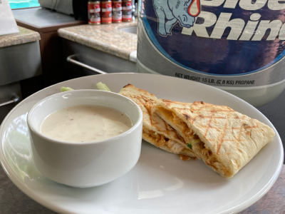 Sandwich cut in half sitting on one side of a round white plate. On the other side of the plate is a dipping bowl with blue cheese dressing. Fresh Blue Rhino propane tank in the background.