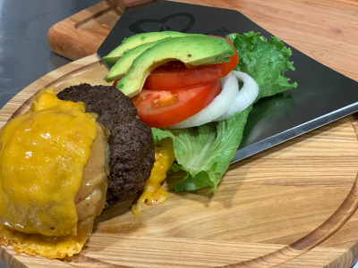On a round wooden cutting board is a bun with a slice of cheese on it, grilled hamburger, slices of tomatoes, onions, avocado, lettuce.