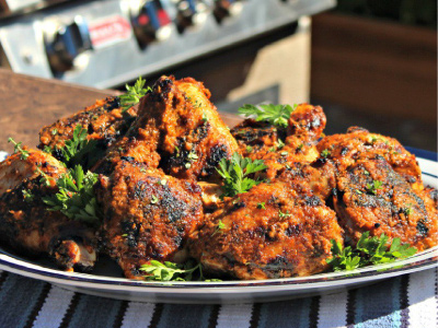 Grilled chicken breasts stacked on a white plate, sitting on a white and blue striped tablecloth with a standup propane grill in the background.