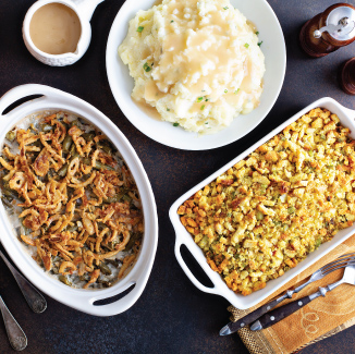 Top view of finished stuffing, green bean casserole, and mash potatoes in white bowls on a black countertop.