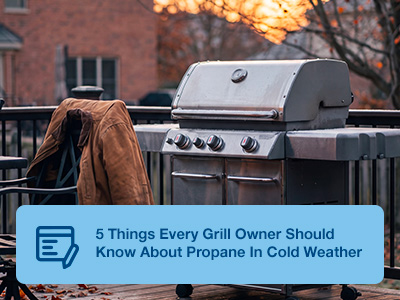 A standup propane grill on a deck with a coat sitting on a chair in front of the grill.
