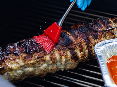 Person applying a marinade to a pork loin with a basting brush, while on a standup propane grill.