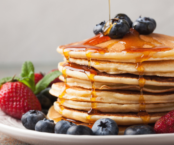 Stack of grilled fluffy pancakes with maple syrup, blueberries, strawberries on a white plate.
