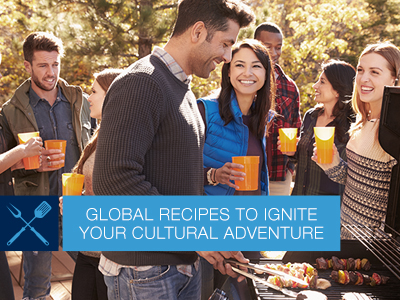 Man in fall attire outdoors on the patio grilling food at a standup gas grill, with other friends in fall attire in the background.