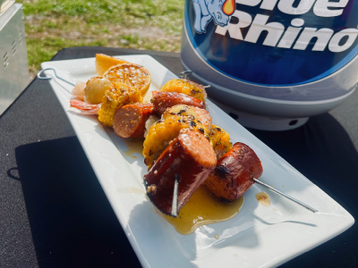 Two finished Louisiana “Low Country” Skewers on a white plate, on a wooden cutting board, with a fresh Blue Rhino and standup propane grill in the background.