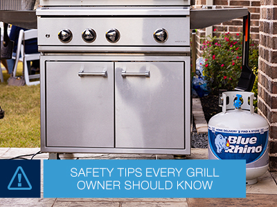 Stainless steel standup propane grill with a Blue Rhino tank sitting next to the grill, sitting on a stone patio in a backyard.