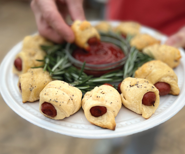 Finished pigs in a blanket on a white plate around rosemary with a bowl of ketchup.