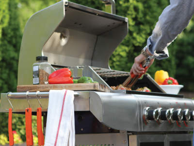Man with plaid long sleeve shirt standing in front of stand up propane grill, using utensil to move food.