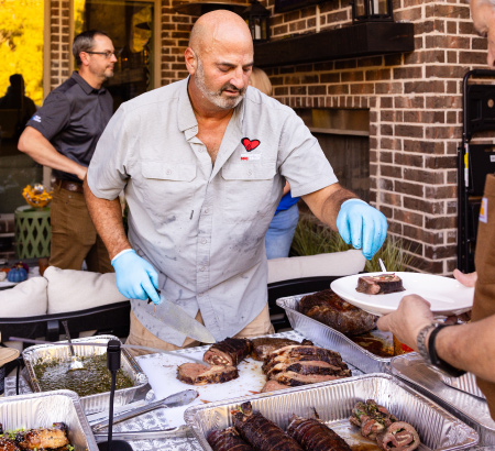 Chef serving grilled slices roasts, over a table of finished grilled food.