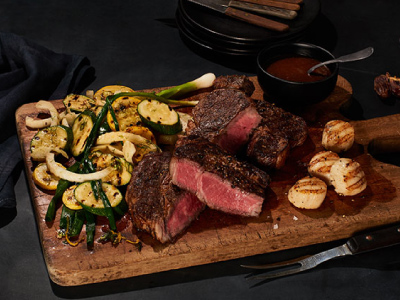 Grilled Ribeye with grilled vegetables and scallops on a wood cutting board with a bowl of A.1 sauce