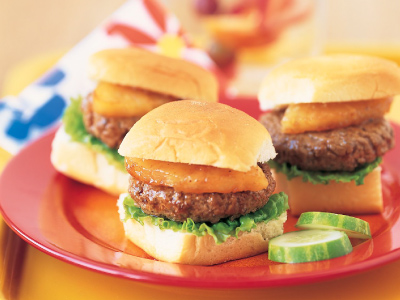 Mini beef parties topped with grilled pineapple and lettuce on Hawaiian sweet rolls with cucumber on a red plate on a yellow table.