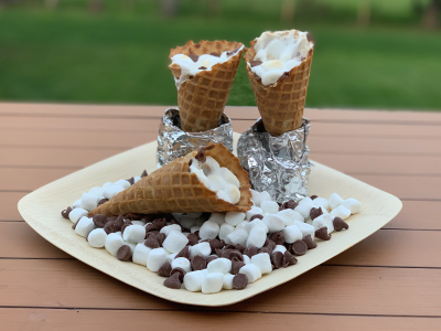 Two finished s'mores waffle cones standing up aluminum foil at the base, with mini marshmallows and chocolate chips and a third s'mores waffle cones on a white plate, sitting on a wood patio table, with grass in the background.