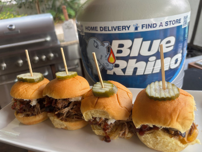 Four finished Southern Style Pulled Pork Sliders on a white plate, with a fresh Blue Rhino tank and standup propane grill in the background.